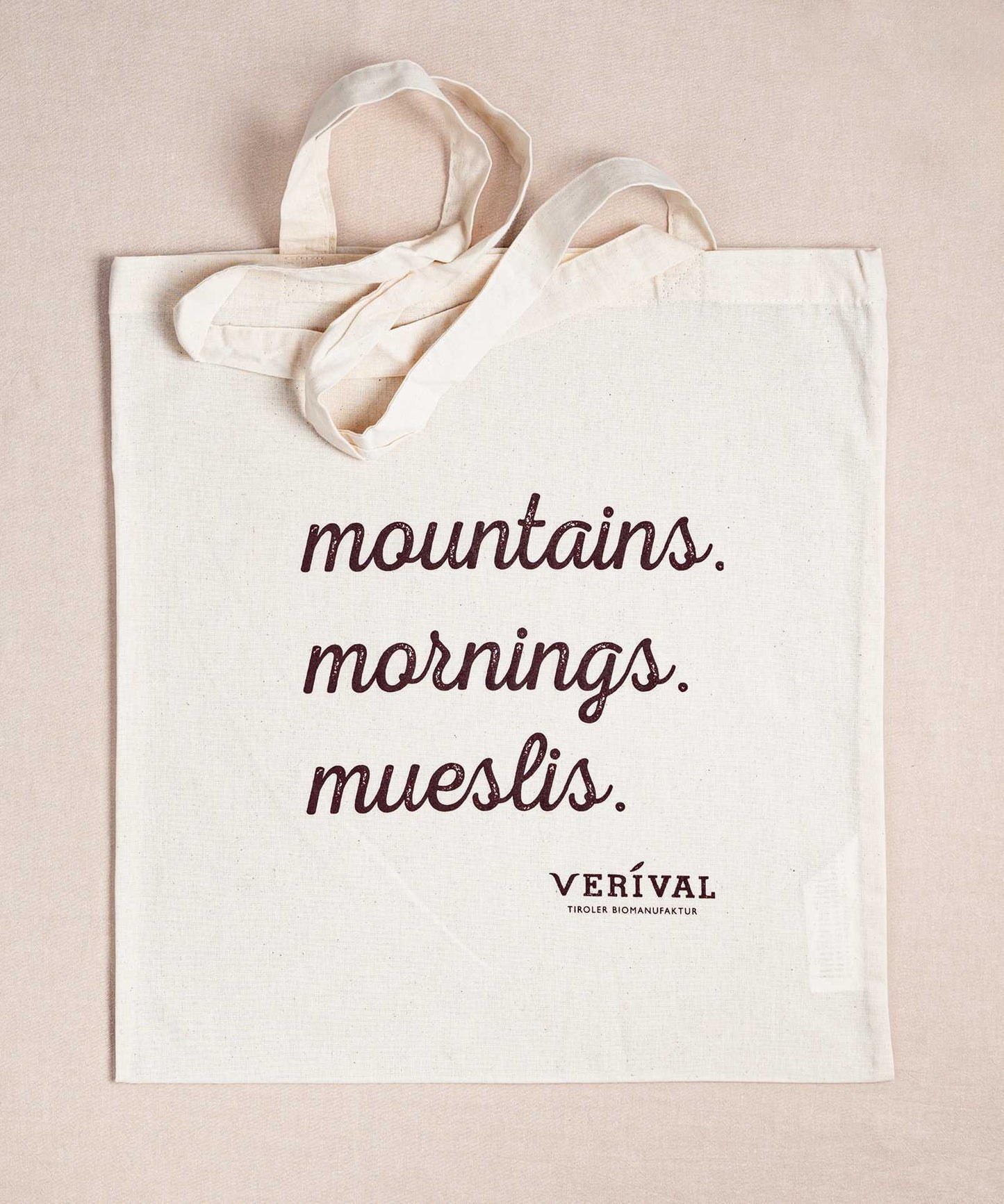Eine beige Baumwolltasche mit Henkeln trägt in dunkelbraunen Buchstaben die Aufschrift „mountains. mornings. mueslis.“ und spricht damit Porridge-Liebhaber an. Darunter steht in kleinerer Schrift „Verival“. Sie liegt flach auf einer passenden Unterlage und ist Teil des Sets "Porridge Lover“ von Verival Bio, ideal für biobewusste Menschen.