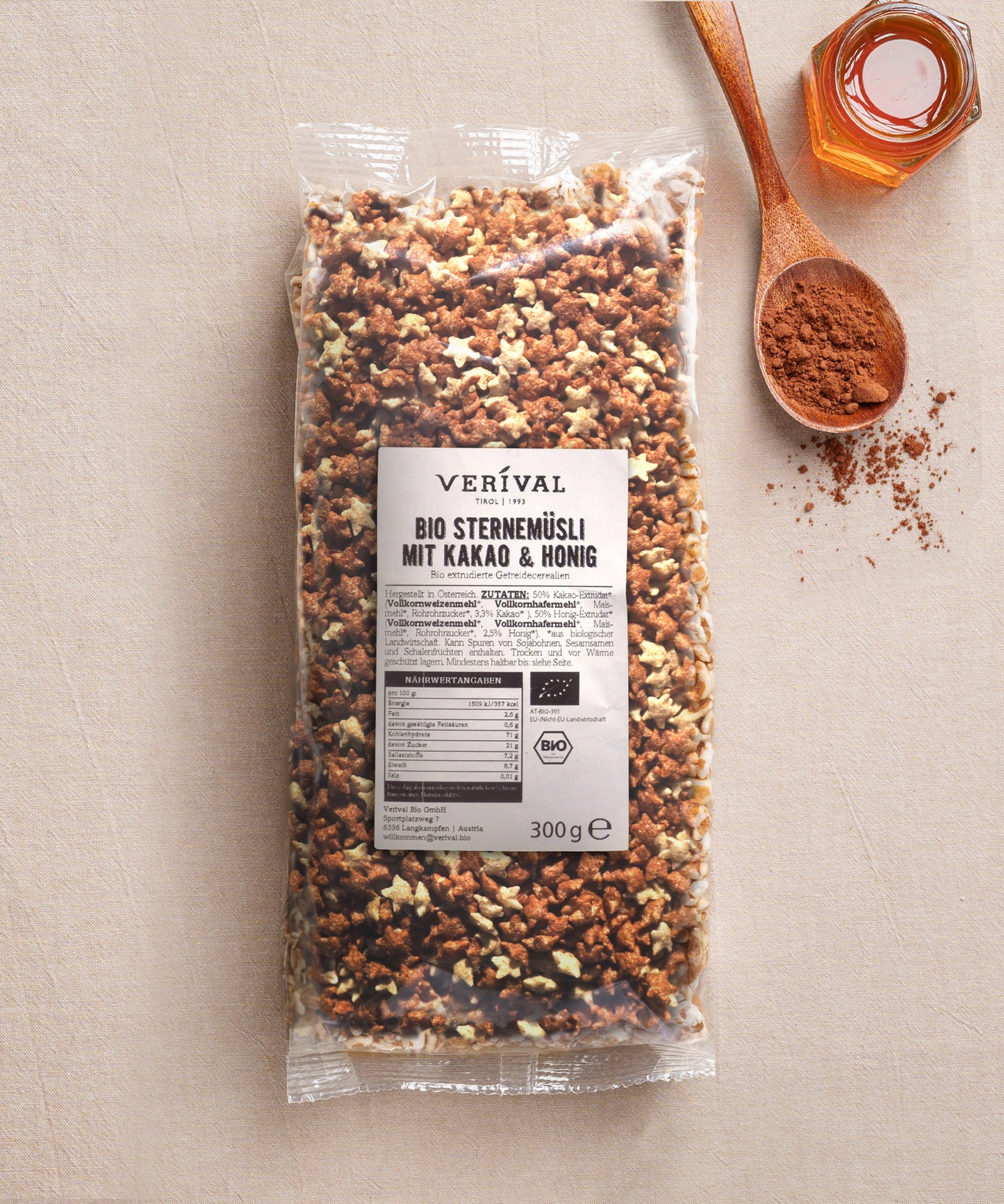 Eine 300g-Packung Verival Bio GmbH Bio Sternemüsli mit Kakao & Honig steht auf einer beigen Fläche, daneben ein Holzlöffel mit Kakaopulver und ein kleines Glas Honig.
