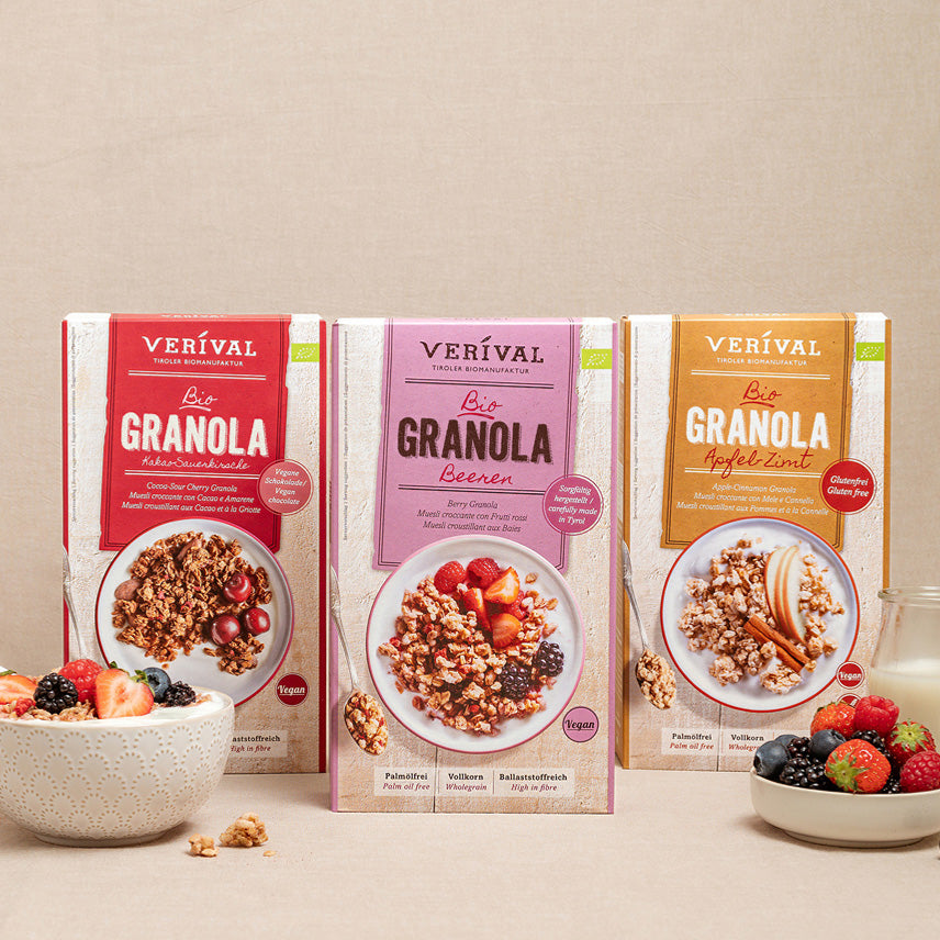 Drei Schachteln Verival-Granola in verschiedenen Geschmacksrichtungen stehen aufrecht auf einer beigen Fläche, davor steht eine Schale mit Granola und gemischten Beeren.