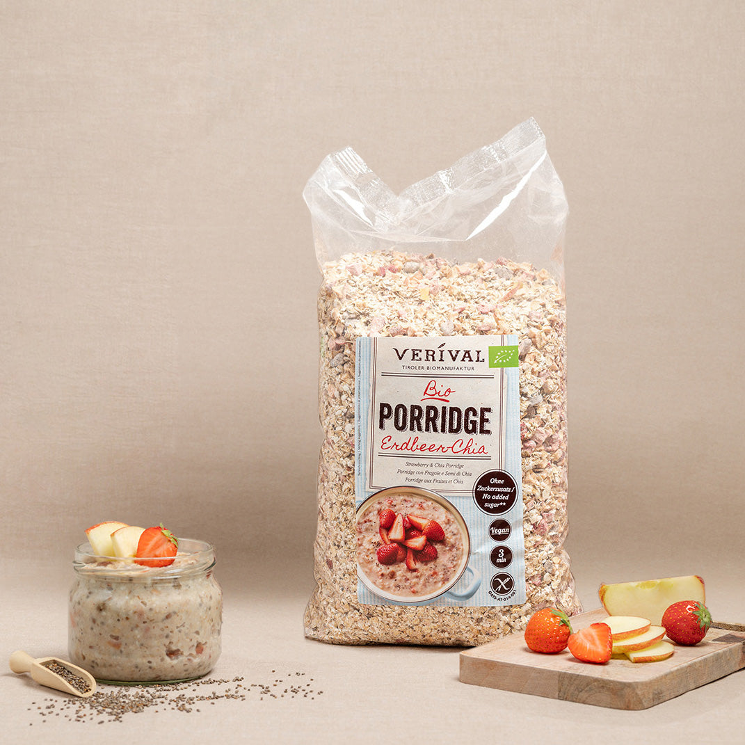 Eine Tüte Verival Bio Porridge Enduleer Chia steht aufrecht neben einer Schüssel mit zubereitetem Porridge mit Obst, ganzen Erdbeeren, Apfelscheiben und einer Kugel Chiasamen auf einem Tisch.