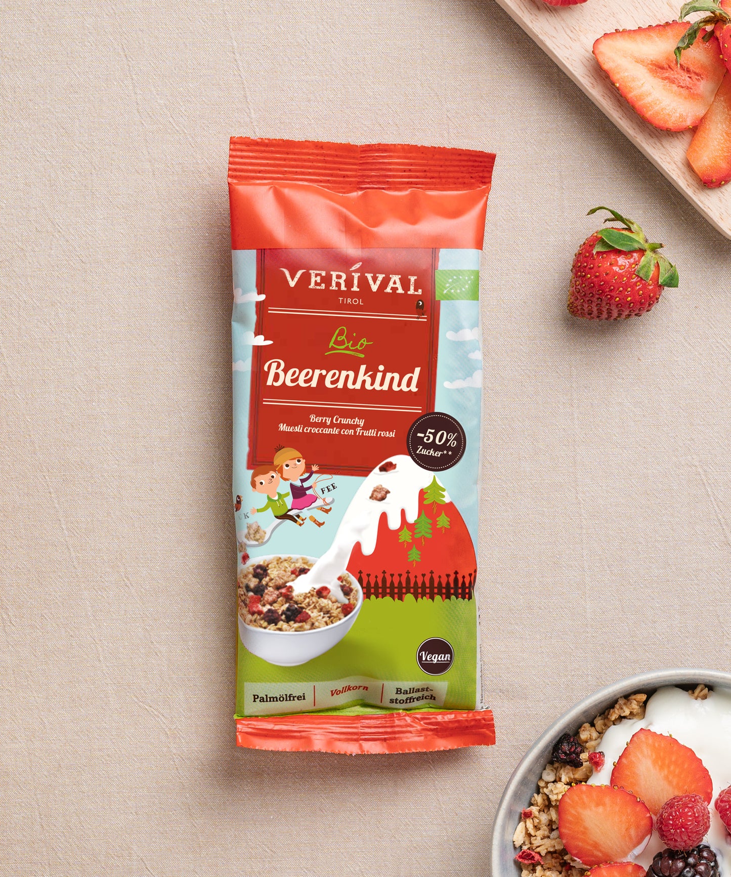 Eine Packung Verival Bio GmbH Bio Beerenkind Beerenmüsli liegt auf einer beigen Fläche neben einer Schale veganem Crunchy Frühstück mit Milch und geschnittenen Erdbeeren.