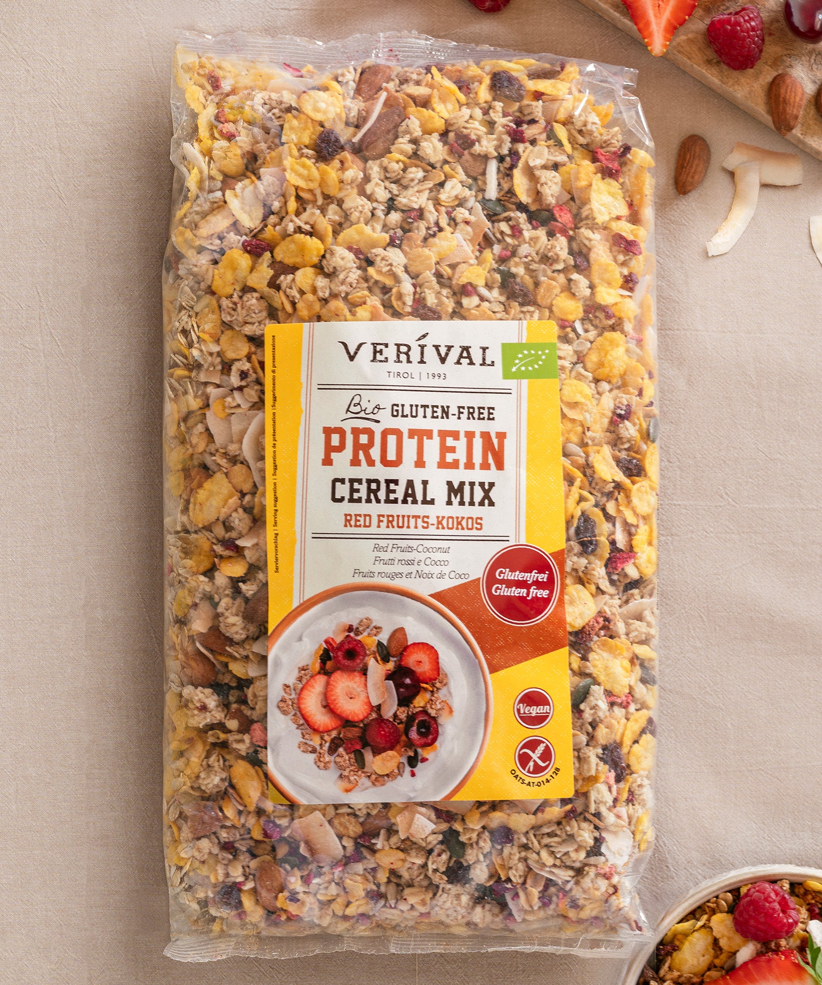 Eine versiegelte Packung Verival Bio GmbH Bio Protein Cereal Mix Red Fruits-Kokos glutenfrei liegt auf einer beigen Fläche, umgeben von verstreuten Nüssen und Früchten; diese ballaststoffreiche Mischung ist perfekt für einen nahrhaften Start in den Tag.