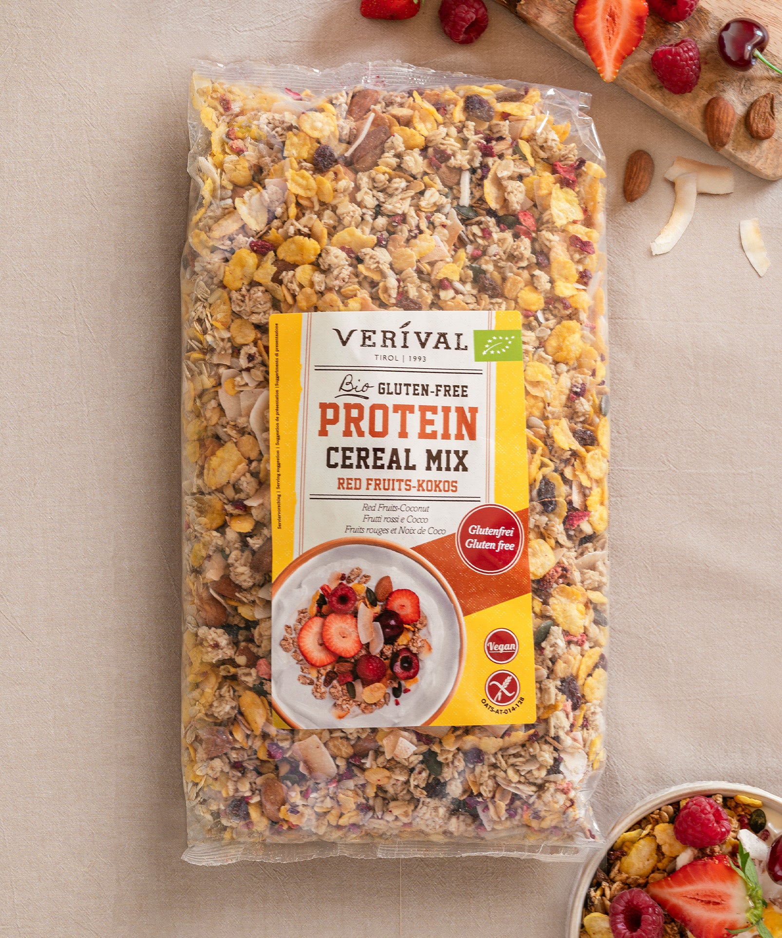 Eine Packung Verival Bio GmbH Bio Protein Cereal Mix Red Fruits-Kokos glutenfrei liegt auf einer beigen Fläche neben einer kleinen Schüssel mit dem ballaststoffreichen Müsli, daneben liegen Früchte und Nüsse.