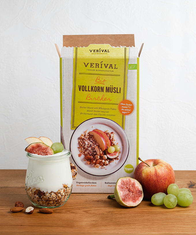Neben einer Packung Verival Bio Bio Bircher Vollkorn-Müsli steht ein Glas, das mit Schichten aus Joghurt, Müsli, Apfelscheiben, Feigen und Weintrauben gefüllt ist, während auf der Holzplatte frisches Obst angerichtet ist.